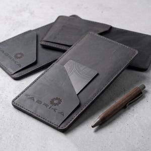 Puede incluir: Varios monederos de cuero gris con la palabra "FABRIKA" grabada y un logotipo. Un monedero muestra una tarjeta en su bolsillo. Un bolígrafo de madera descansa junto a los monederos.