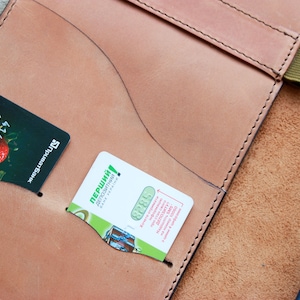 May include: A brown leather notebook cover with a green elastic band closure. The cover has two pockets for cards, one with a credit card and the other with a business card. The business card has the text "перший!" in green.