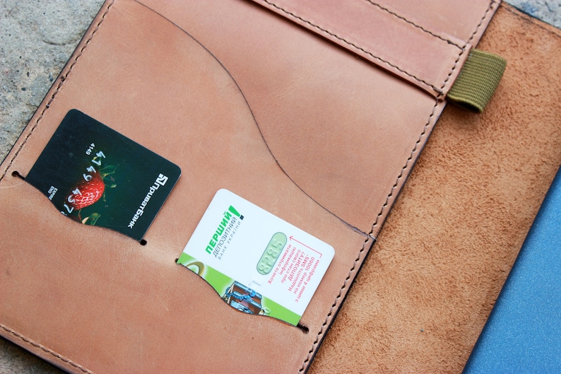 May include: A brown leather notebook cover with a green elastic band closure. The cover has two pockets for cards, one with a credit card and the other with a business card. The business card has the text "перший!" in green.