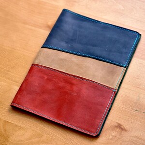 Peut inclure: Une couverture de carnet en cuir avec trois panneaux de couleurs différentes : rouge, beige et bleu. Les bords sont cousus avec un fil bleu clair.