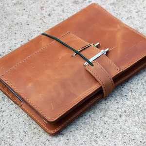 Könnte beinhalten: Ein braunes Leder-Notizbuchcover mit einem Riemen und einem Stifthalter. Die Abdeckung ist geschlossen und hat einen abgenutzten Look.