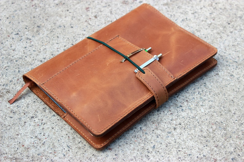 K&ouml;nnte beinhalten: Ein braunes Lederjournal mit Stifthalter und gr&uuml;nem Gummibandverschluss. Das Journal hat ein Lesezeichen und einen Riemenverschluss. Der Stift ist silber und gr&uuml;n. Das Journal ist ca. 20 cm lang und 15 cm breit.