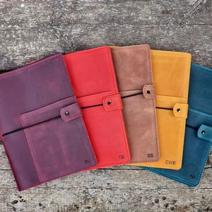 May include: Five leather-bound journals in different colours, including burgundy, red, brown, yellow, and blue. Each journal has a black elastic closure and a small pocket on the front cover. The journals are labelled with initials: "RN", "TB", "SS", "CWB", and "TP".