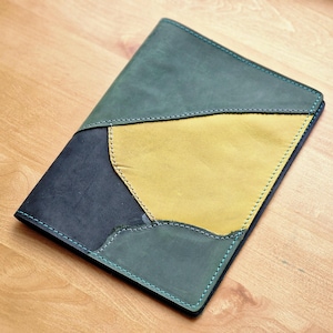 May include: A handmade leather notebook cover with a patchwork design. The cover features sections of teal, navy, and yellow leather, stitched together with teal thread. The notebook cover is rectangular and lies on a wooden surface.