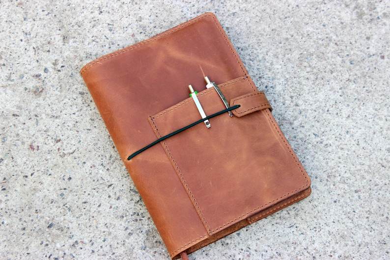 K&ouml;nnte beinhalten: Ein braunes Lederjournal mit Stifthalter und schwarzem Gummibandverschluss. Zwei Stifte sind in den Stifthalter eingesetzt. Das Journal liegt auf einer Betonoberfl&auml;che. Das Leder hat ein Used-Look-Aussehen.
