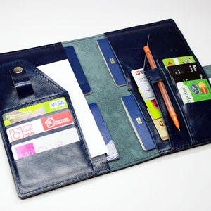 Puede incluir: Cartera de viaje de cuero azul marino abierta con múltiples ranuras para tarjetas, bolsillos para pasaporte y un portalápices. La cartera está abierta, revelando varias tarjetas de crédito, un bolígrafo y un pasaporte.
