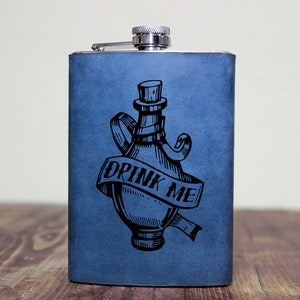 May include: A blue stainless steel flask with a black and white illustration of a bottle with a cork and a ribbon that says "Drink Me".