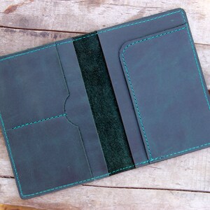 May include: A dark green leather passport holder with a teal stitching detail. The holder has multiple pockets for cards and cash.