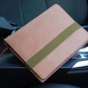 May include: A light brown leather notebook with a green elastic band closure. The notebook is sitting in the center console of a car.
