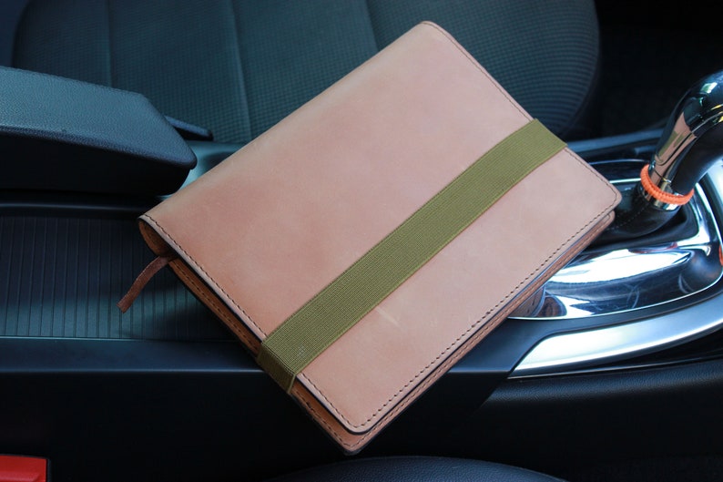 May include: A light brown leather notebook with a green elastic band closure. The notebook is sitting in the center console of a car.