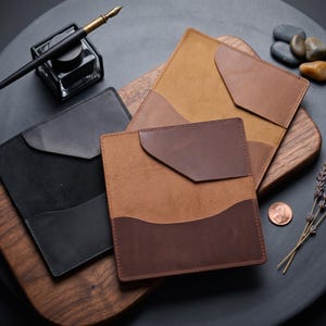 May include: An overhead shot displays several leather wallets in various shades of brown and black, arranged on a wooden surface. A black inkwell with a pen, a penny, and sprigs of lavender are also present, suggesting a stationery or office theme.