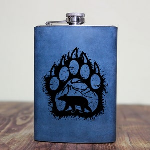 May include: A blue stainless steel flask with a black bear paw print design. The paw print features a silhouette of a bear inside the print.
