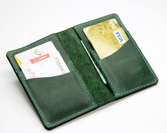 Peut inclure: Un portefeuille en cuir vert avec deux fentes pour cartes et une poche pour l'argent. Le portefeuille est ouvert et montre deux cartes à l'intérieur.