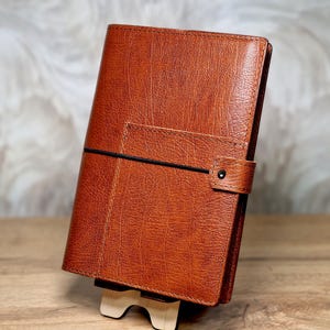 May include: A brown leather-bound journal with a black elastic closure and a pocket on the front cover.
