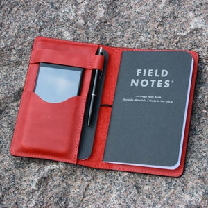 May include: A red leather notebook cover with a phone pocket and pen holder. The cover is open to reveal a black Field Notes notebook with the text "FIELD NOTES" and "48-Page Note Book Durable Materials / Made in the U.S.A."