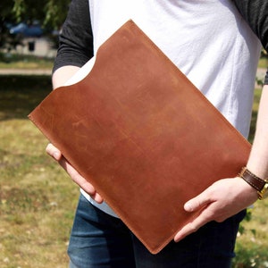 Puede incluir: Una funda para portátil de cuero marrón con cierre de solapa. La funda está siendo sostenida por una persona.