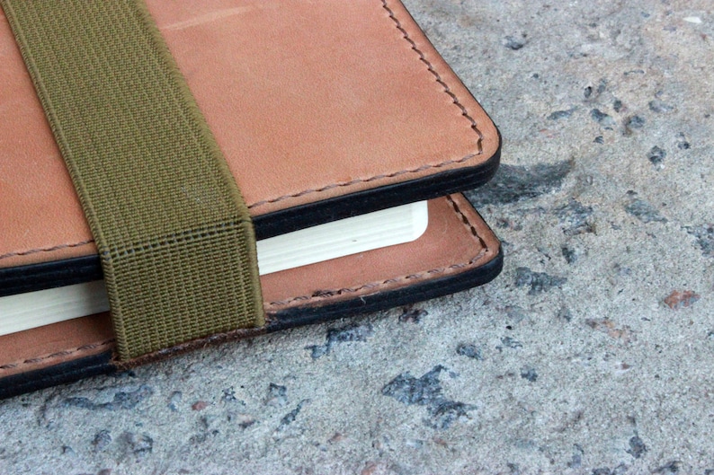 May include: A brown leather notebook with a green elastic band closure. The notebook has a black stitched edge and a white page showing.