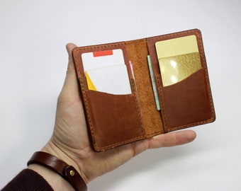 Peut inclure: Portefeuille en cuir marron ouvert, révélant plusieurs fentes pour cartes contenant diverses cartes. Le portefeuille est maintenu ouvert, mettant en valeur son intérieur et ses coutures. Le cuir a un grain naturel et une teinte chaleureuse.