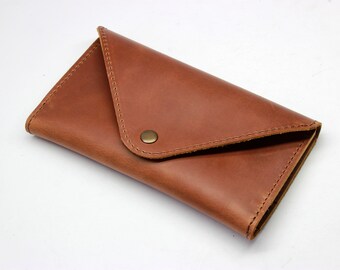 Puede incluir: Una cartera de cuero marrón con cierre de solapa y un broche de latón. La cartera es rectangular y parece hecha a mano. El cuero tiene un grano natural y un tono cálido y terroso.