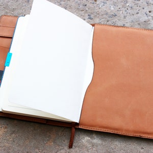 May include: A brown leather bound journal with a blank white page open. The journal has a blue ribbon bookmark and a card tucked into the side pocket.