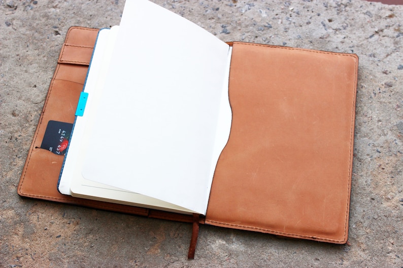 May include: A brown leather bound journal with a blank white page open. The journal has a blue ribbon bookmark and a card tucked into the side pocket.