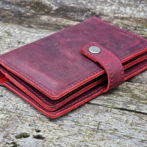 May include: A burgundy leather wallet with a flap closure and a silver button. The wallet is folded and lying on a wooden surface.
