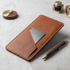 Könnte beinhalten: Ein brauner Lederkartenhalter mit teilweise eingelegter Karte. Ein Holzstift mit Messingakzenten, eine kleine Messing- und Holzscheibe, ein Tintenfass und eine Uhr befinden sich ebenfalls auf der Oberfläche.