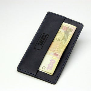May include: A dark blue leather wallet with a blue stitch detail and a money clip. The wallet has the text "Faktor Goldsu" embossed on the front. A 100-ruble note is visible in the money clip.