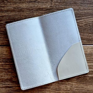 May include: A pale grey leather passport holder with a pocket on the right. The holder is open, showing the inside. The edges are stitched with white thread. It rests on a wooden surface.