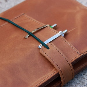 K&ouml;nnte beinhalten: Ein braunes Lederjournal mit Riemenverschluss und Stifthalter. Das Journal wird mit einem dunkelgr&uuml;nen Gummiband und einem silbernen Stiftclip gesichert. Das Leder hat ein strukturiertes Aussehen.