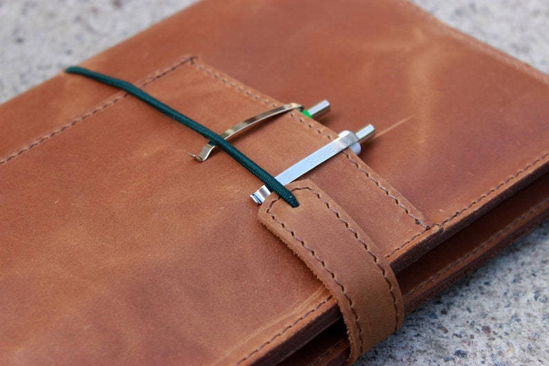 K&ouml;nnte beinhalten: Ein braunes Lederjournal mit Riemenverschluss und Stifthalter. Das Journal wird mit einem dunkelgr&uuml;nen Gummiband und einem silbernen Stiftclip gesichert. Das Leder hat ein strukturiertes Aussehen.