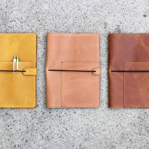 K&ouml;nnte beinhalten: Drei Lederjournale in Gelb-, Beige- und Braunt&ouml;nen werden gezeigt. Jedes Journal hat eine Tasche, einen schwarzen elastischen Verschluss und einen Stifthalter. Die Journale sind rechteckig und scheinen handgefertigt zu sein.