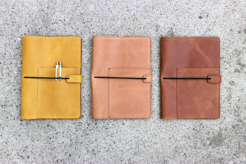 K&ouml;nnte beinhalten: Drei Lederjournale in Gelb-, Beige- und Braunt&ouml;nen werden gezeigt. Jedes Journal hat eine Tasche, einen schwarzen elastischen Verschluss und einen Stifthalter. Die Journale sind rechteckig und scheinen handgefertigt zu sein.