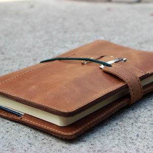 K&ouml;nnte beinhalten: Ein braunes Lederjournal mit einem Stift, der mit einem gr&uuml;nen Gummiband und einem Lederriemen befestigt ist. Das Journal ist geschlossen, wobei die Seiten am Rand sichtbar sind. Das Journal liegt auf einer Betonoberfl&auml;che.