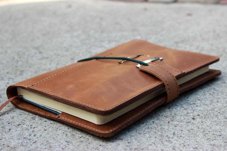 K&ouml;nnte beinhalten: Ein braunes Lederjournal mit einem Stift, der mit einem gr&uuml;nen Gummiband und einem Lederriemen befestigt ist. Das Journal ist geschlossen, wobei die Seiten am Rand sichtbar sind. Das Journal liegt auf einer Betonoberfl&auml;che.