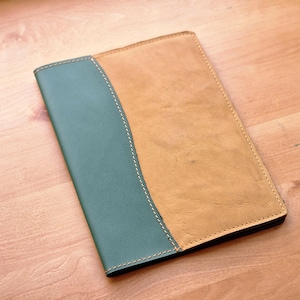 May include: A leather journal with a two-tone design. The left side is teal, and the right side is tan. The two sections are separated by a curved seam with white stitching. The journal is closed and lying on a wooden surface.