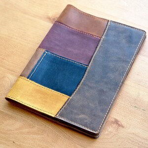 May include: A leather notebook cover with a patchwork design. The cover is made of four different colors of leather: brown, purple, blue, and yellow. The edges of the cover are stitched together.