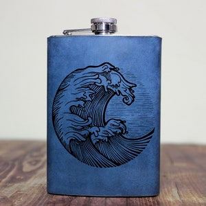 May include: A blue leather flask with a silver top and a black and white engraved image of a wave.