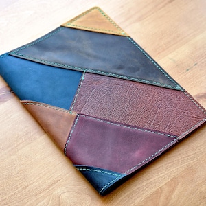 May include: A handmade leather case with a patchwork design. The case features various colors, including brown, blue, and burgundy, stitched together with green thread. The leather has a textured appearance, and the case is likely for a tablet or notebook.