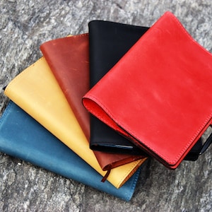 Op de afbeelding: Een stapel van vijf lederen notitieboekjes in verschillende kleuren, waaronder rood, geel, blauw, bruin en zwart. De notitieboekjes zijn in een waaier vorm gerangschikt, met het rode notitieboekje bovenaan. De notitieboekjes zijn allemaal gesloten en hebben een eenvoudig, elegant ontwerp.