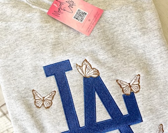 May include: A light gray sweatshirt featuring the letters "LA" in blue, embroidered with butterflies. The butterflies have brown and white wings. A pink tag with the text "Magical Crafts" is attached.