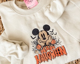 May include: A light beige sweatshirt featuring a Mickey Mouse Halloween design. Mickey Mouse is wearing an orange pumpkin costume and the text "HALLOWEEN" is dripping in orange.