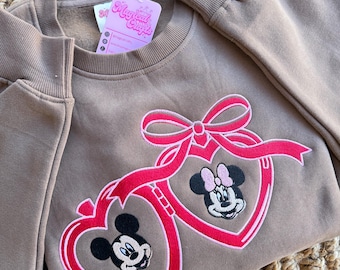 May include: A brown sweatshirt with a pink and red embroidered design of Mickey Mouse and Minnie Mouse in heart-shaped frames. The sweatshirt has a crew neck and long sleeves. The tag on the sweatshirt says "Magical Crafts".