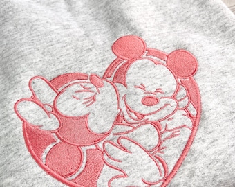 May include: A light gray sweatshirt featuring an embroidered design of Mickey and Minnie Mouse embracing within a pink heart. The sweatshirt has a crew neck and long sleeves. The tag reads "Magical Crafts".