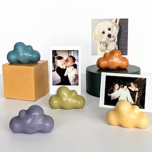 May include: A selection of cloud-shaped ornaments in shades of blue, green, purple, and yellow. Some clouds are displayed with framed photographs. One cloud sits on an orange cube, and another on a dark green cylinder. The photographs are in polaroid style.