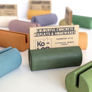 Puede incluir: Una colección de coloridos expositores de hormigón en varios tonos, incluyendo verde, azul y naranja. Cada soporte tiene una ranura para sujetar tarjetas u objetos pequeños. Hecho a mano en el Reino Unido, con el texto "COLOURFUL CONCRETE DISPLAYS & HOMEWARES."