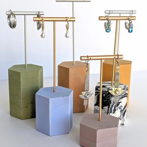 May include: Earring display stands with hexagonal bases in shades of green, blue, brown, and pink. The stands have gold or silver metal posts, showcasing various earrings, including hoops and drop earrings. The bases are made of concrete.