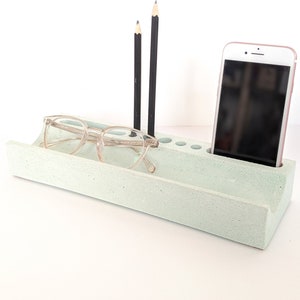 May include: A light green concrete desk organiser with a phone holder, a glasses holder, and slots for pens and pencils.