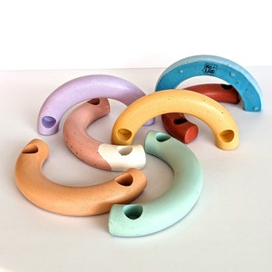 May include: A set of six colourful concrete arches with holes. The arches are in shades of purple, pink, yellow, orange, blue, and green. The arches are arranged in a semi-circle, with the largest arch at the top and the smallest arch at the bottom. The arches are made by Ko-Lab.