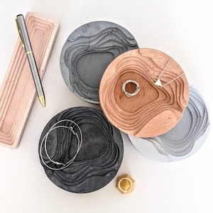 以下が含まれることがあります： グレー、ライトグレー、ブラウン、ブラックの4つのコンクリート製のリングディッシュ。波打つようなテクスチャのある表面をしています。ディッシュは白い表面にペンとジュエリーと一緒に配置されています。